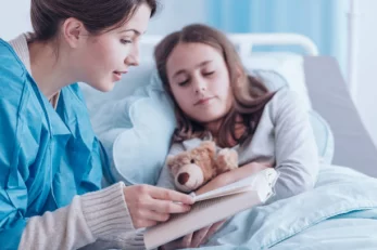 Older Sister Reading Book To Unhealthy Girl With Plush Toy In Private Clinic